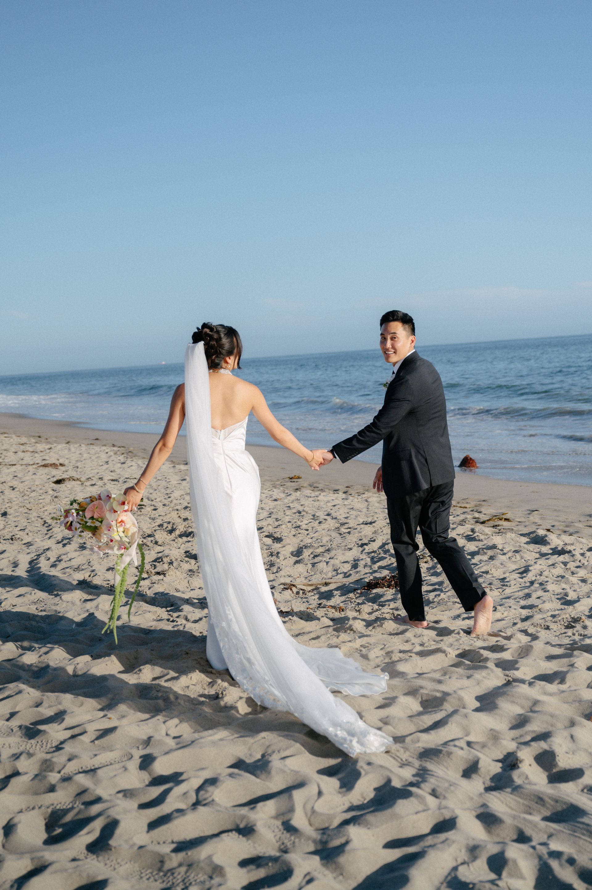 Bride and groom holding hands on a beach, walking toward the ocean at sunset