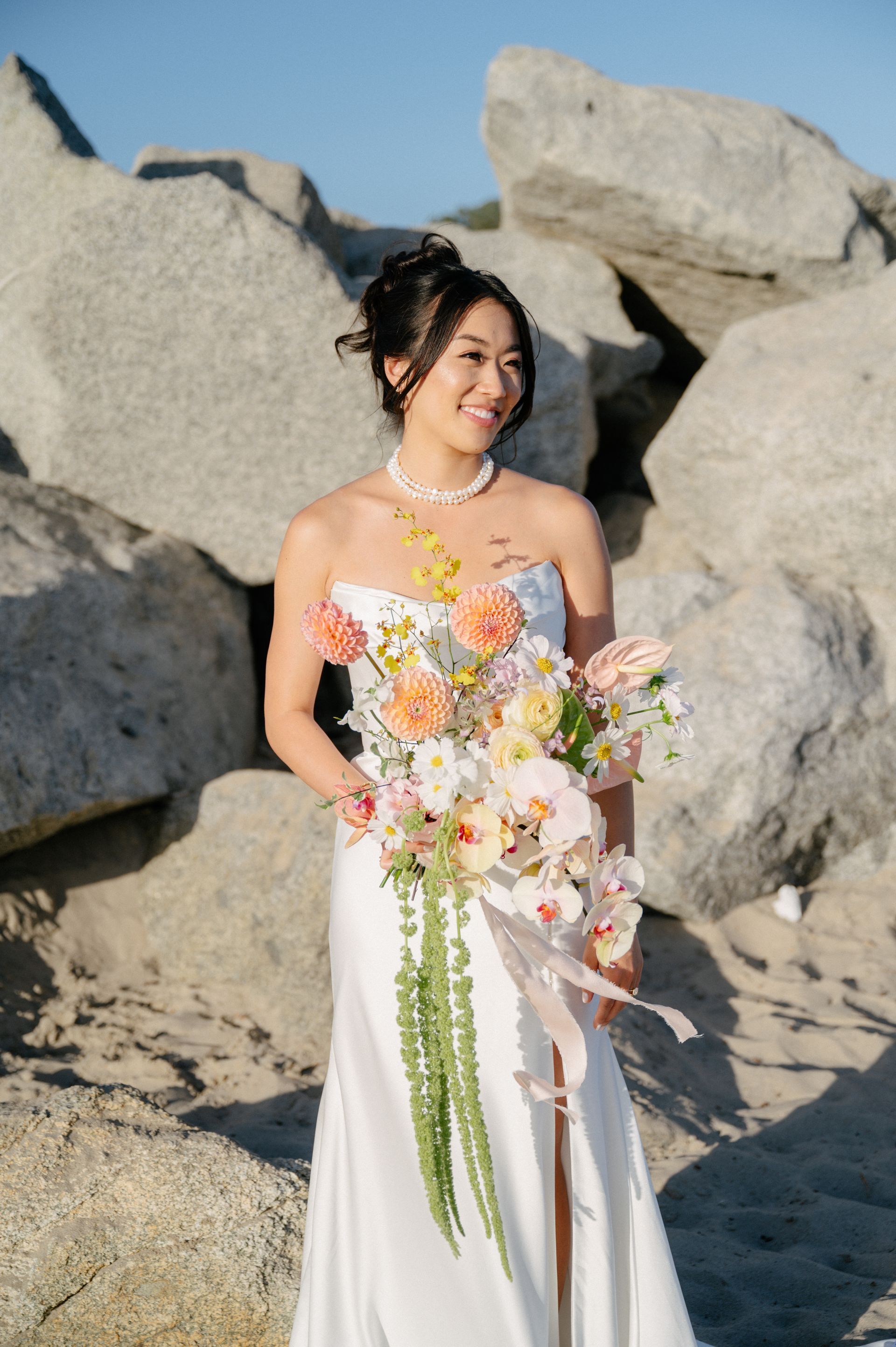 Person in a white dress holding a bouquet, standing on rocky beach cliffs in sunlight.
