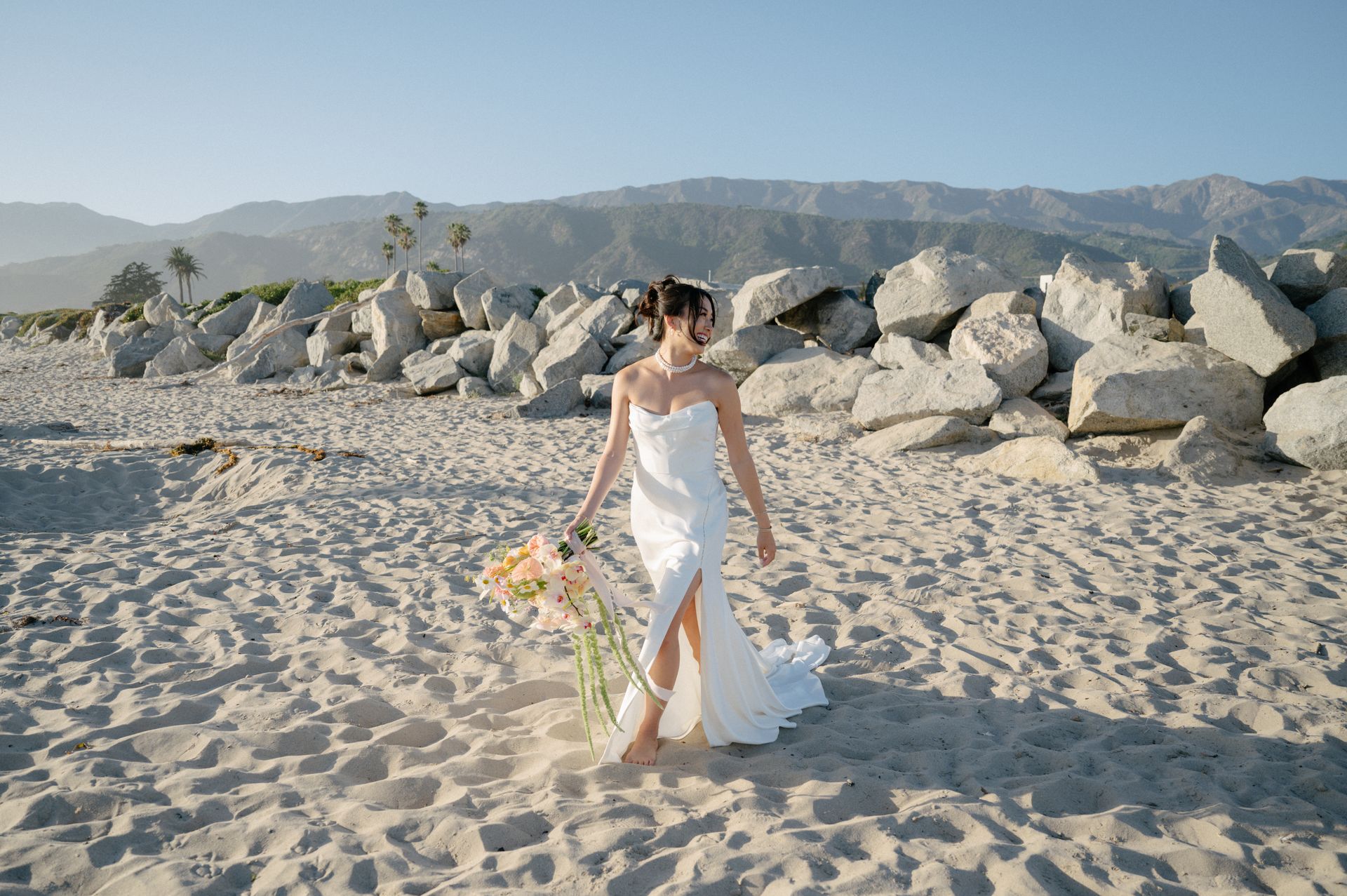 Woman in a white dress walking on a sandy beach beside large rocks, with mountains in the background.