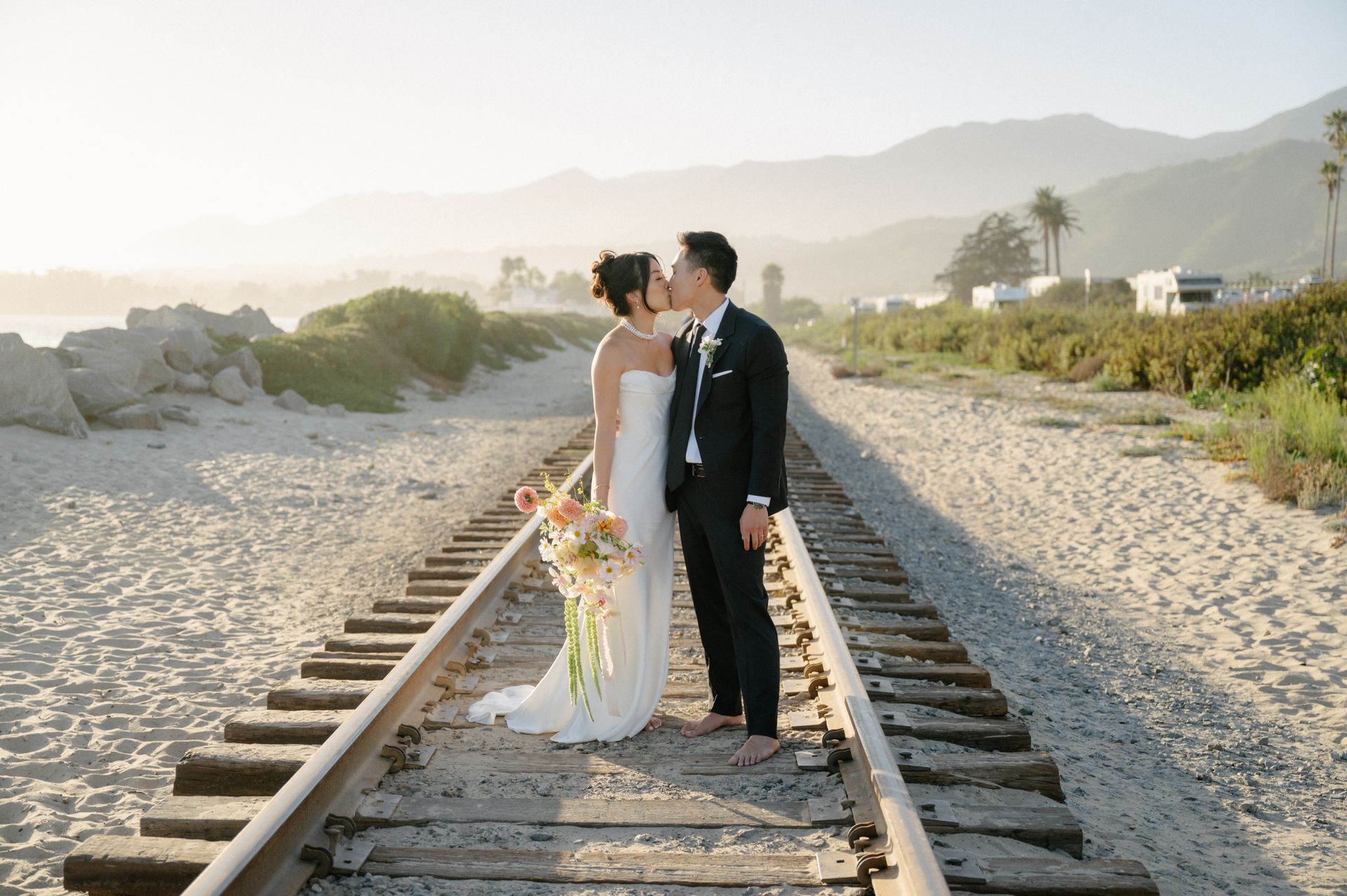 Bride and groom kissing on railroad tracks at sunset with mountains in the background