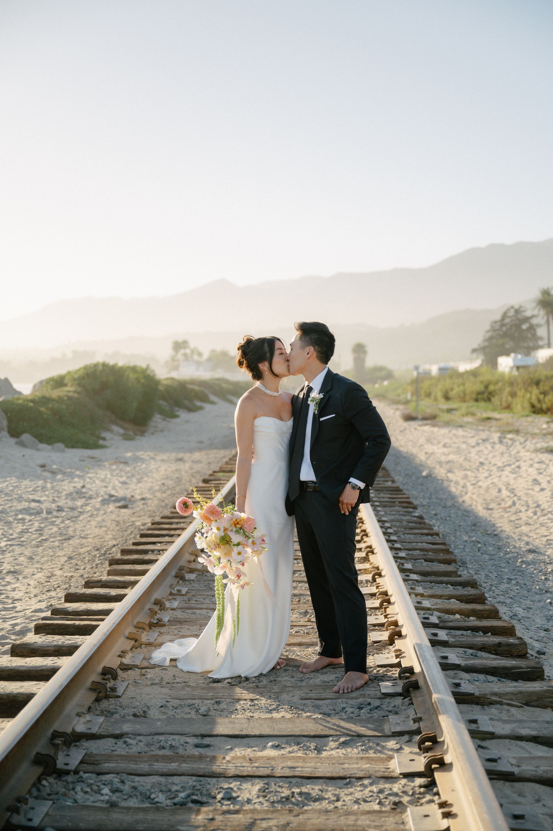 Couple in formal attire kissing on railroad tracks at sunset, with mountains in the background.