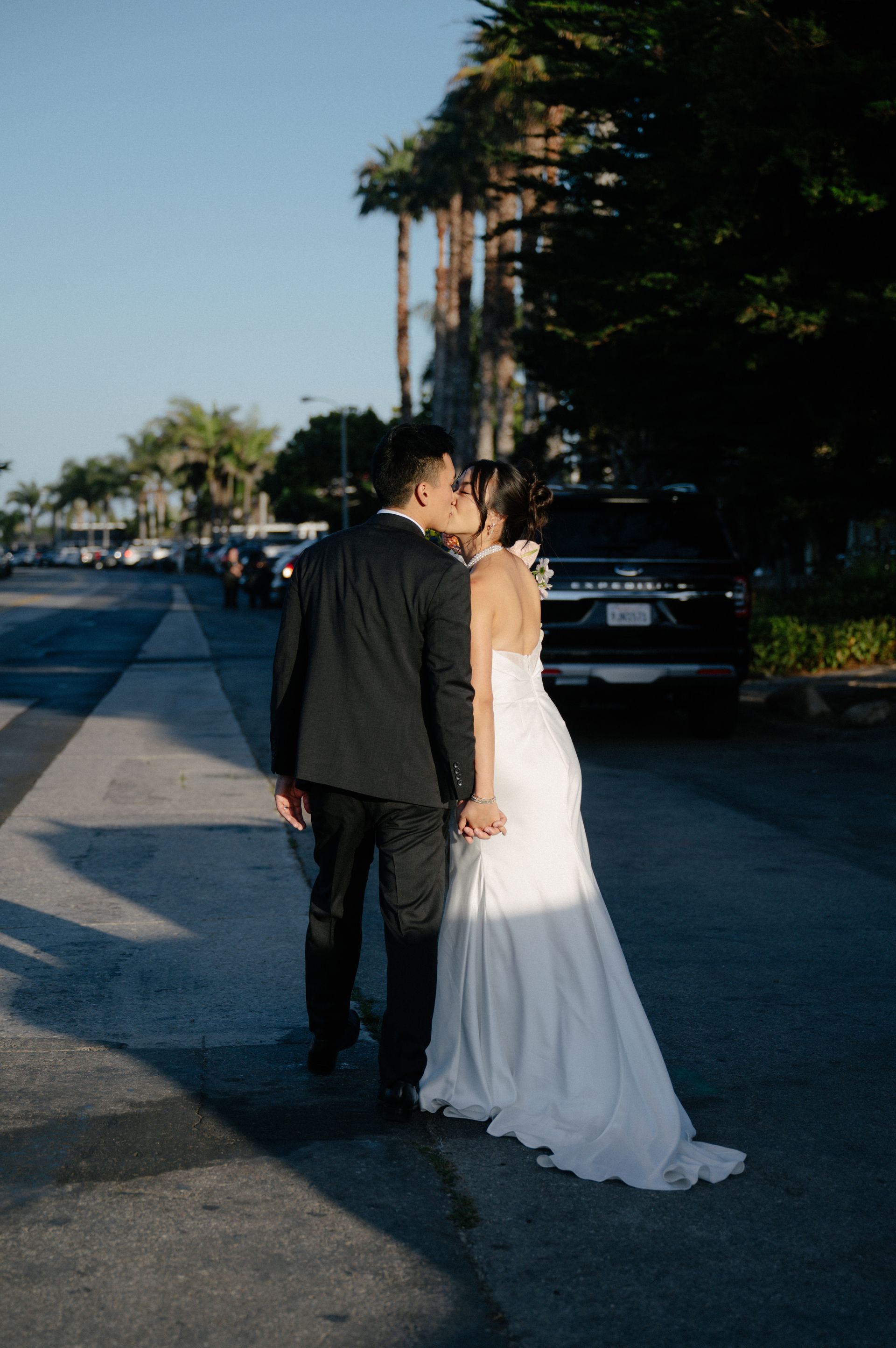 Couple in wedding attire standing on a sunlit street, sharing a kiss near parked cars and tall trees