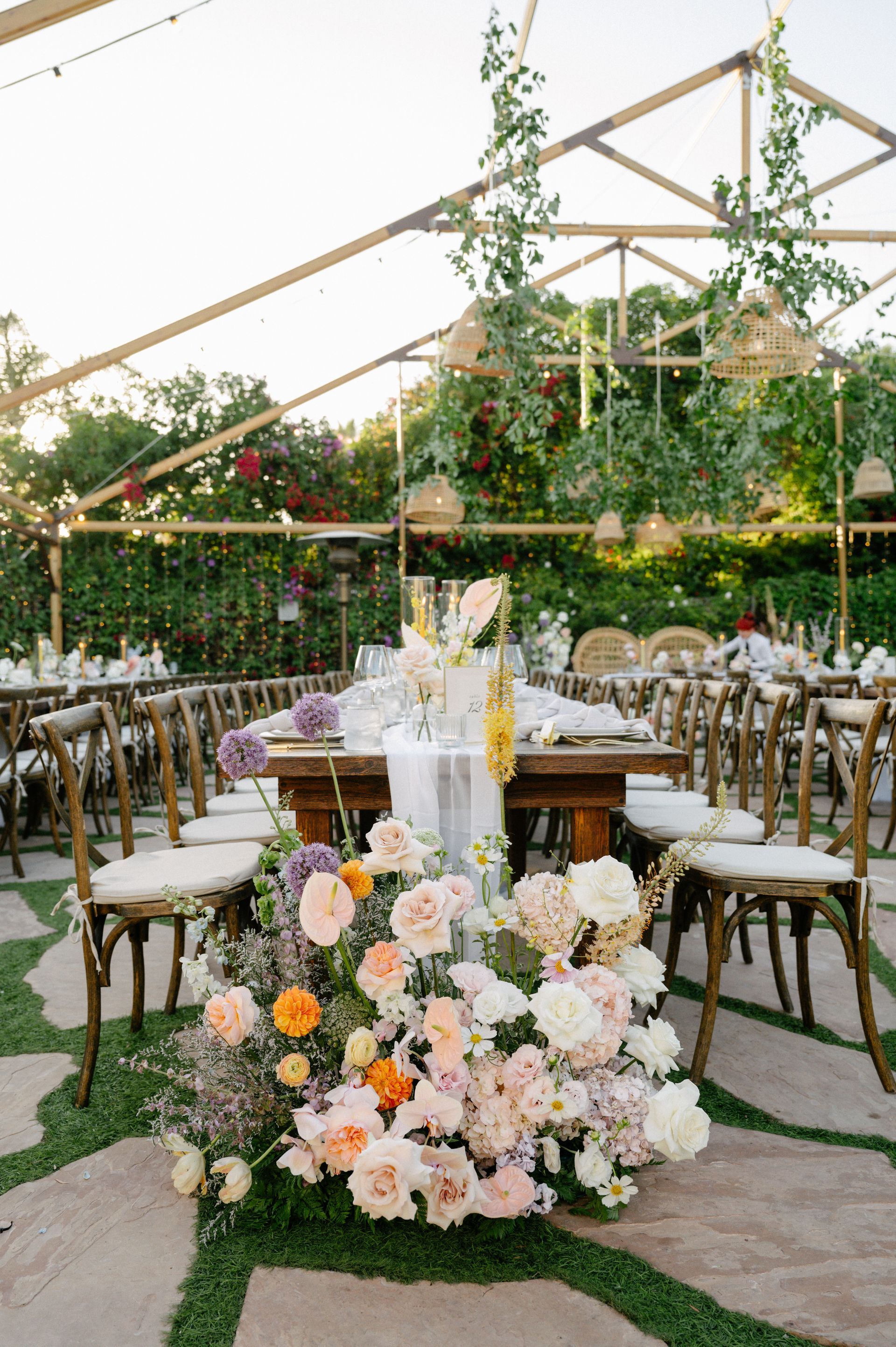 Outdoor wedding reception tent with floral centerpiece, round tables, and hanging greenery decorations