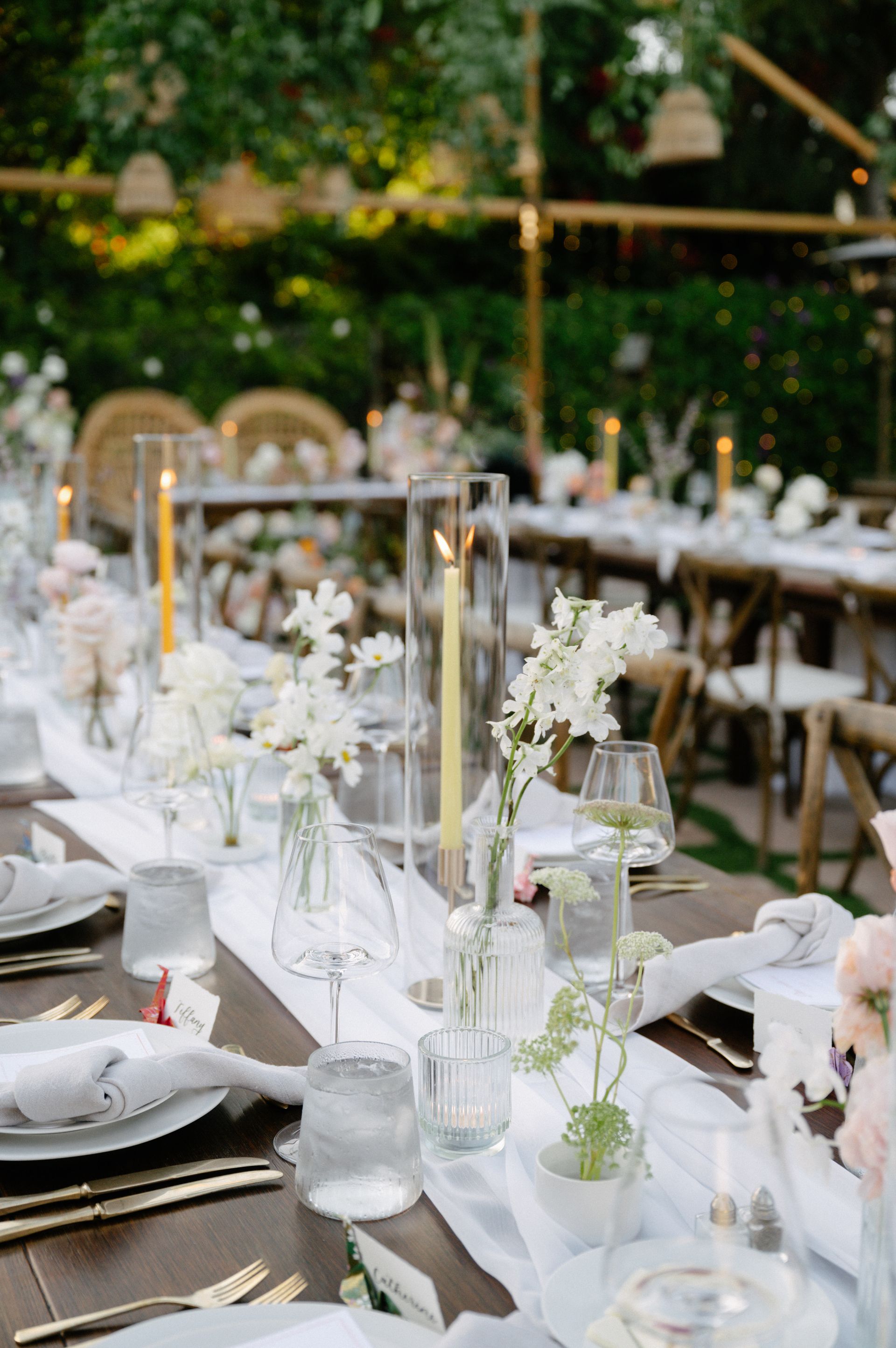 Elegant outdoor dining table set with white flowers, candles, and glassware in a garden setting