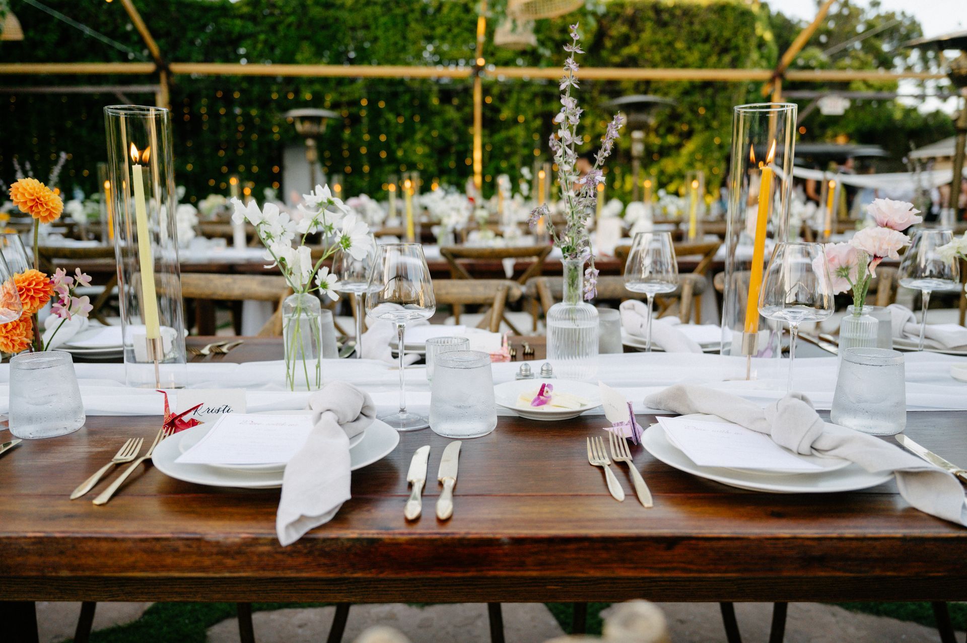 Elegant outdoor banquet table set with white linens, glassware, and floral centerpieces in a garden setting