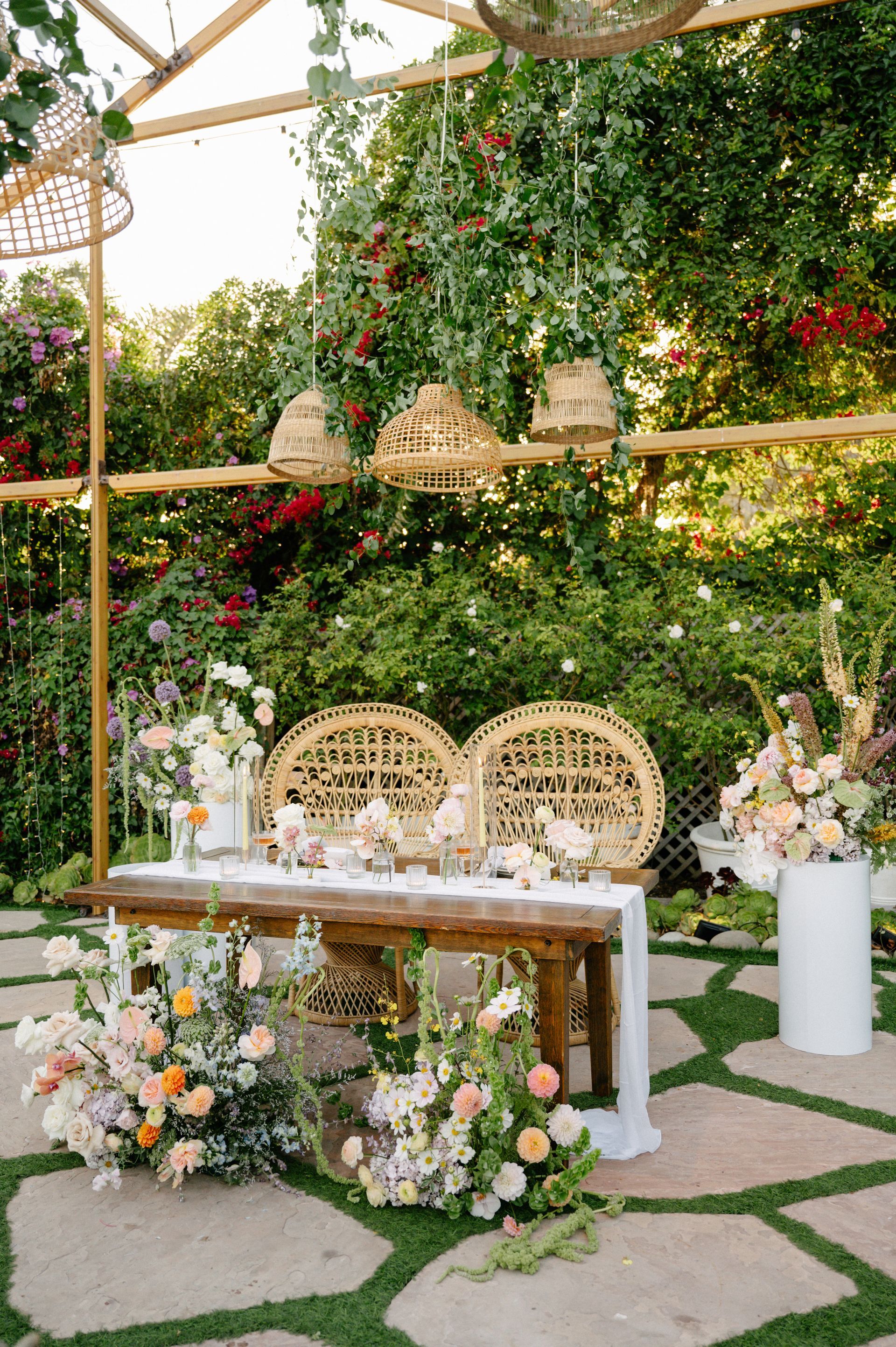 Elegant outdoor wedding table with floral arrangements, wicker chairs, and lush greenery backdrop