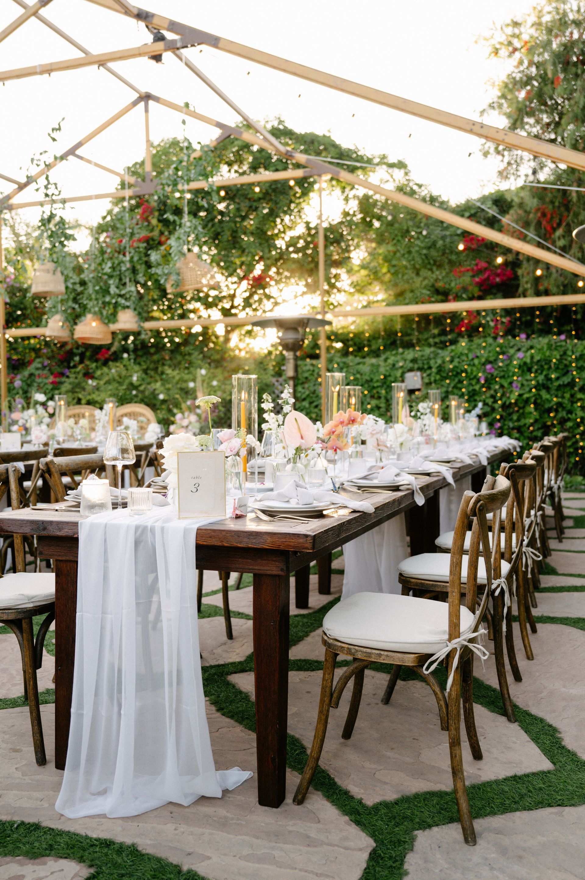 Elegant outdoor dining setup under a tent with long wooden tables, white chairs, and lush floral greenery.
