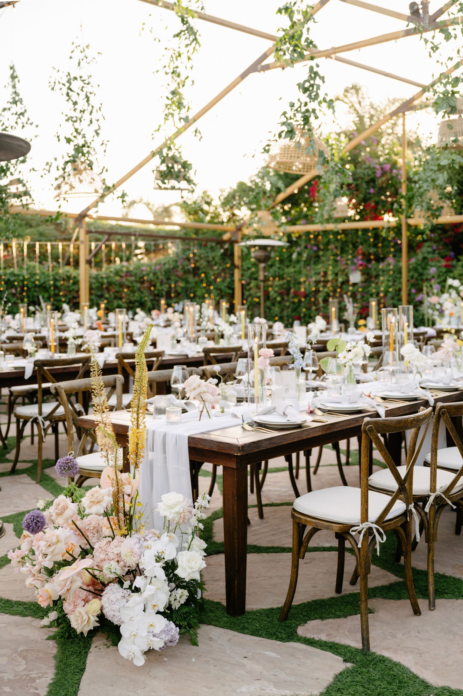 Outdoor wedding reception setup with long wooden tables, white chairs, and floral arrangements under a glass canopy