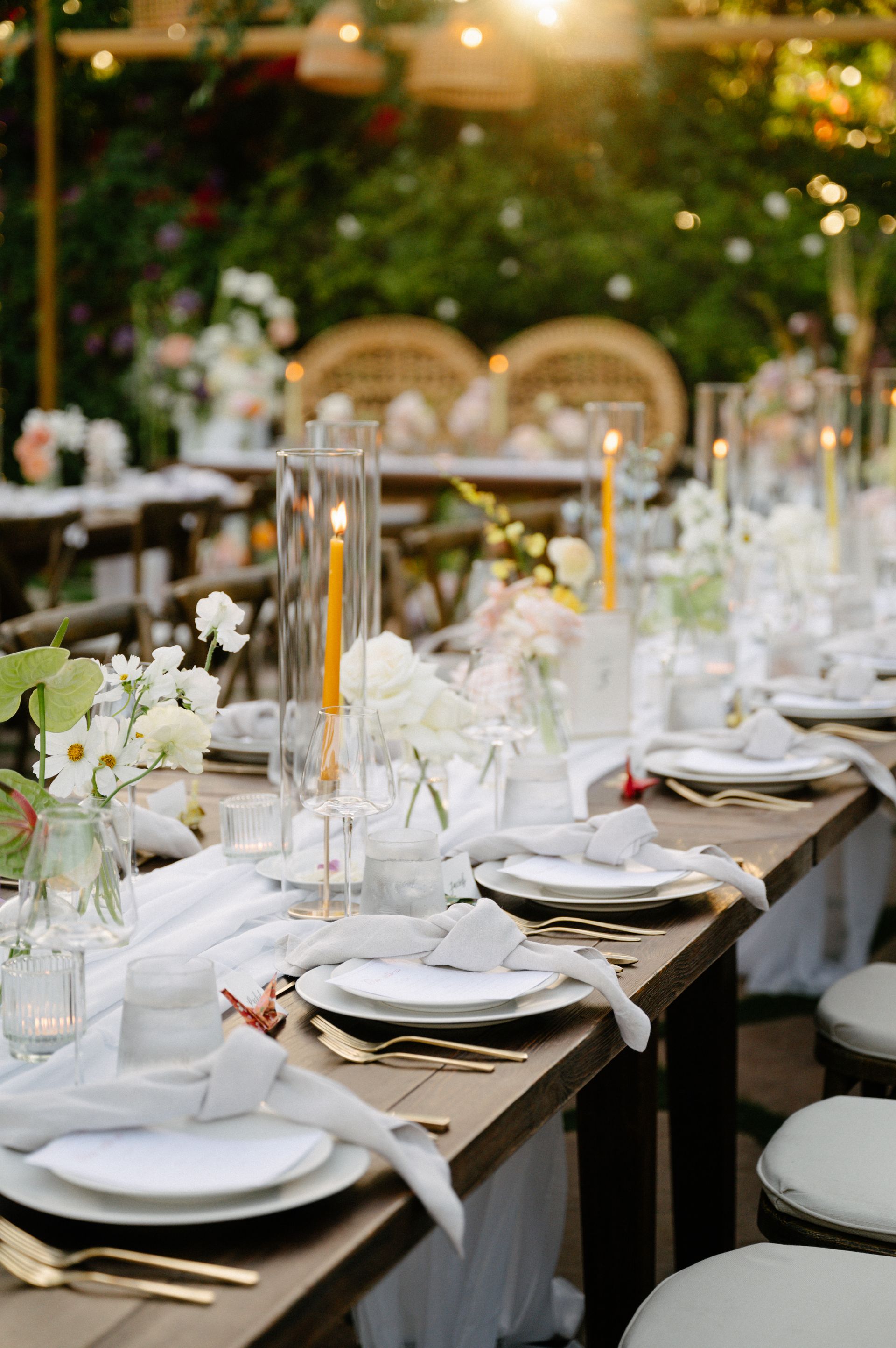 Elegant outdoor dining table set with white plates, candles, and floral centerpieces at dusk