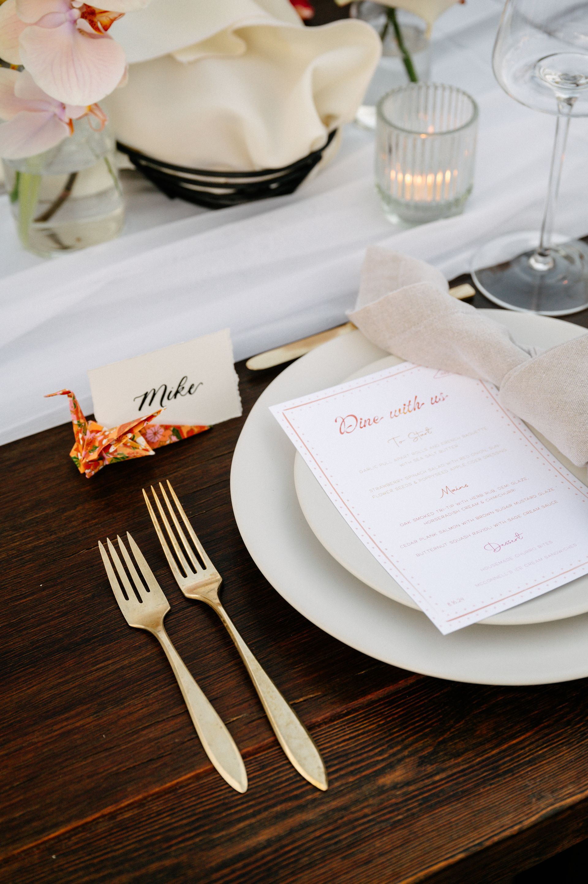 Elegant table setting with white plates, gold flatware, candles, and a menu card on a dark wood table