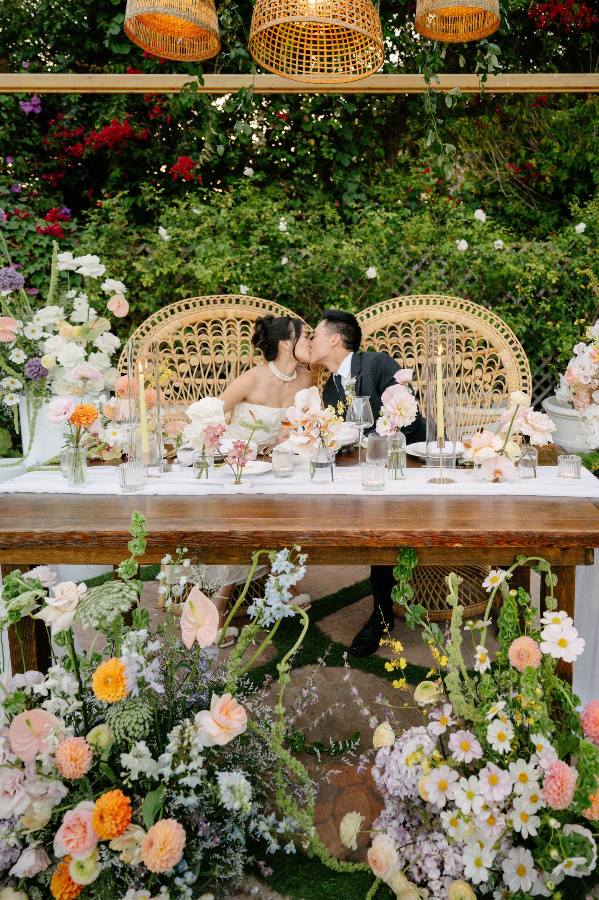 Couple kissing at a floral wedding table with wicker chairs and lush green backdrop