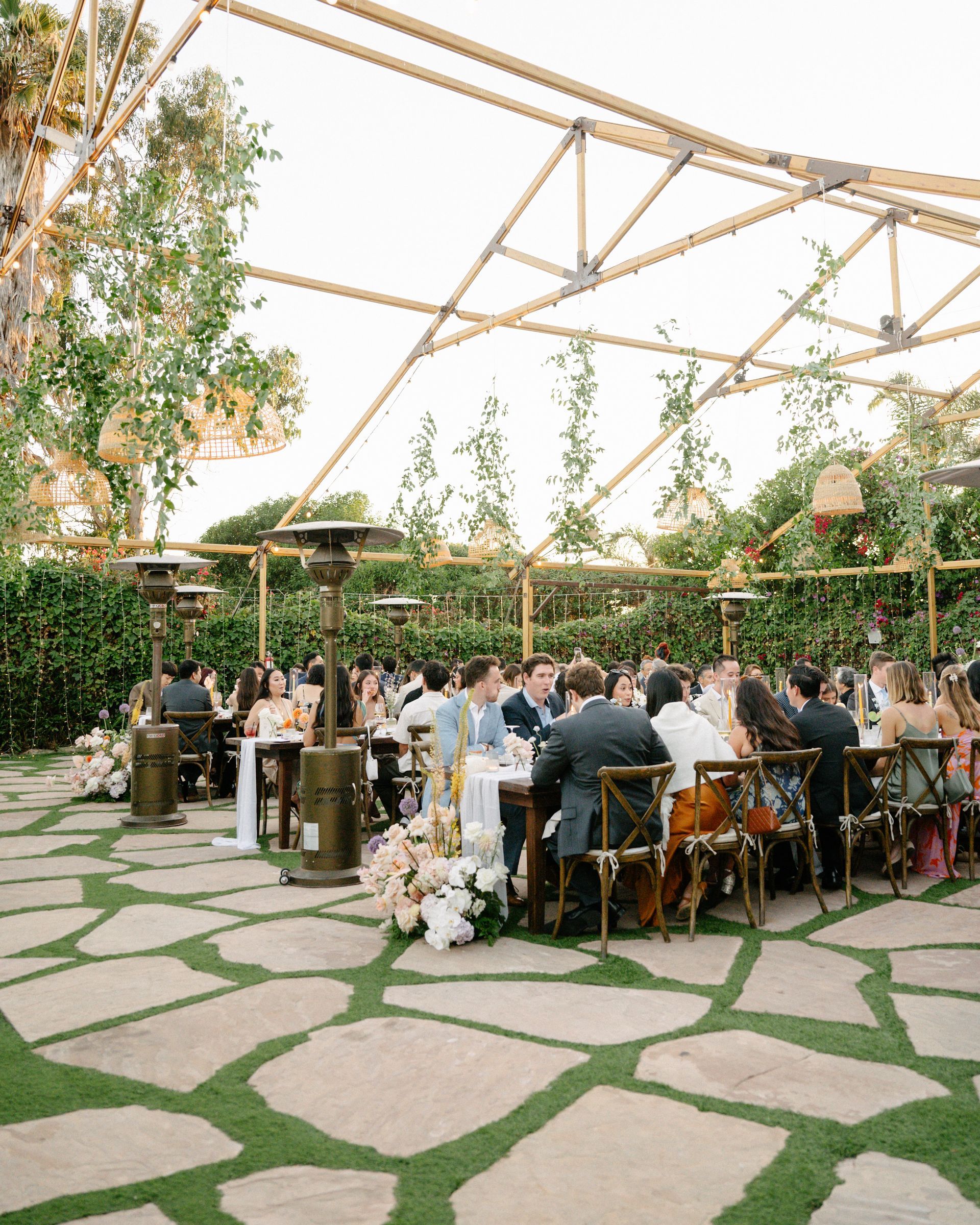 Outdoor wedding reception under a canopy with guests seated at long tables in a garden setting