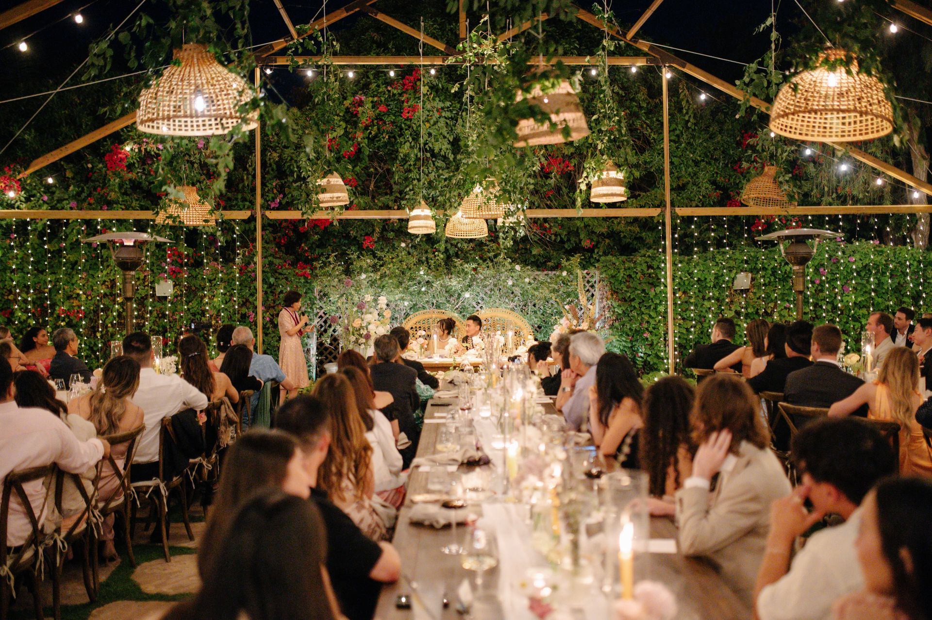 Long banquet table at a warmly lit outdoor wedding reception with guests and hanging greenery lanterns