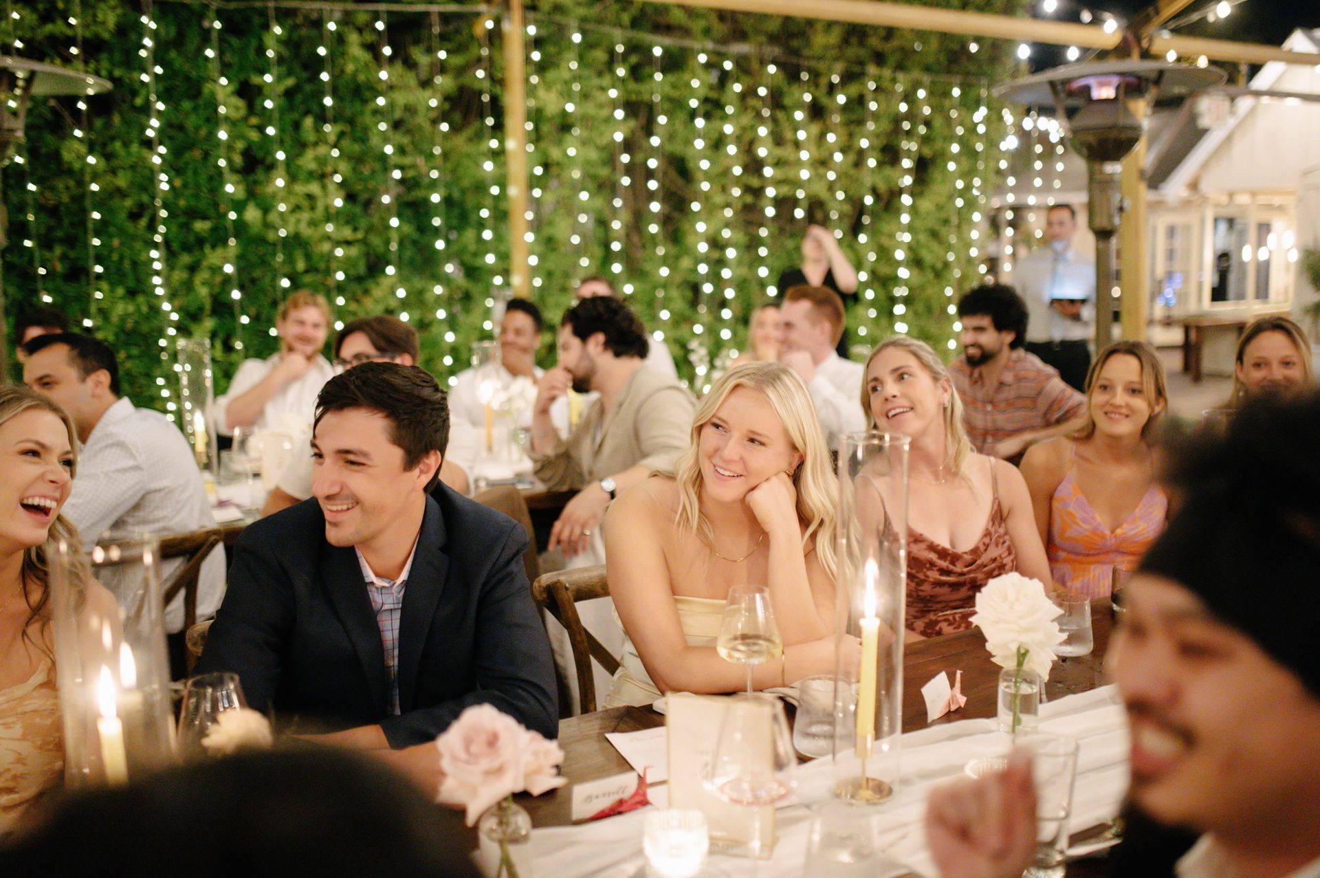 Guests dining at a warmly lit outdoor evening reception with string lights and candles