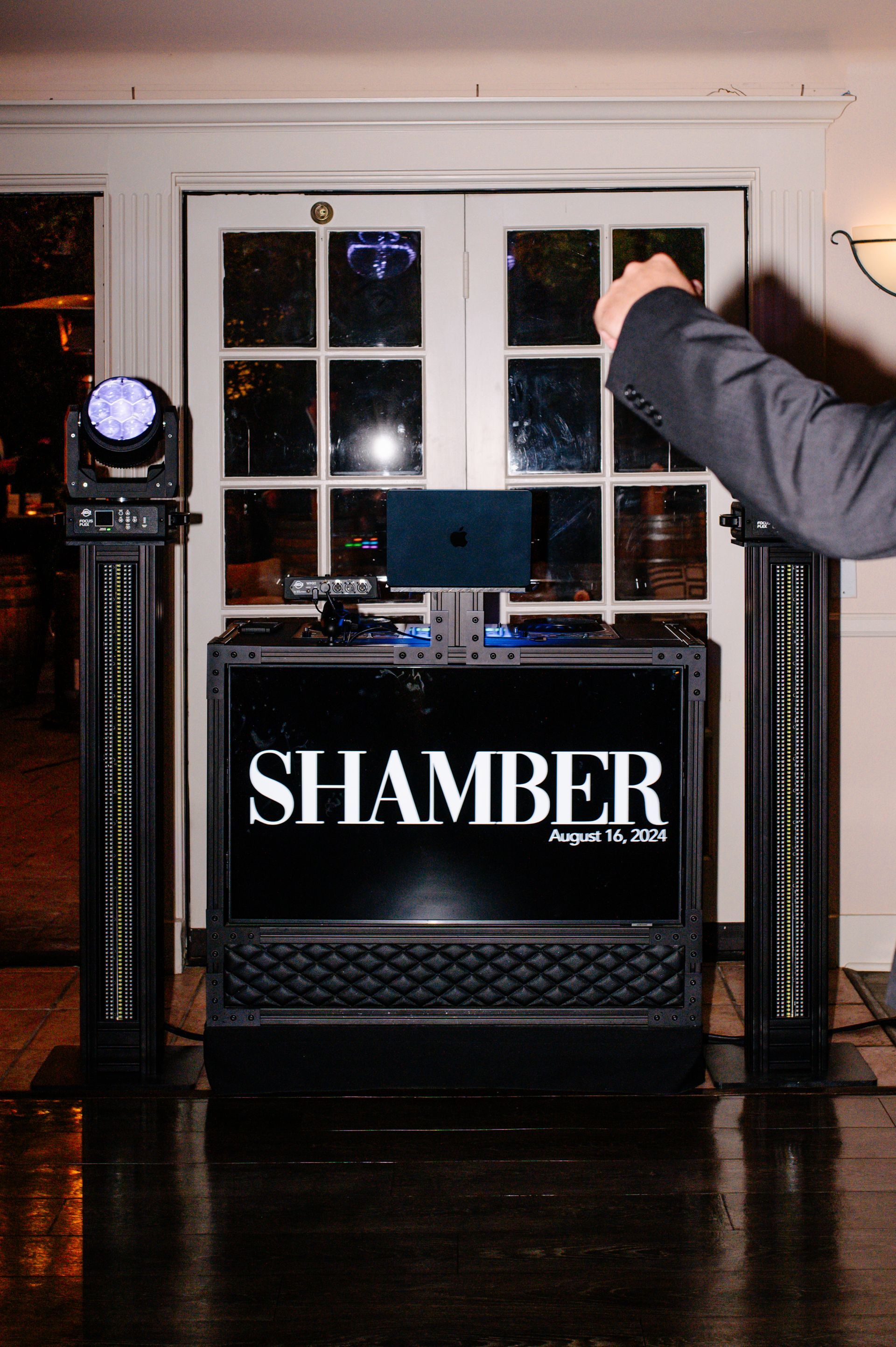 DJ booth with “SHAMBER” sign and a person adjusting equipment in a dimly lit indoor venue