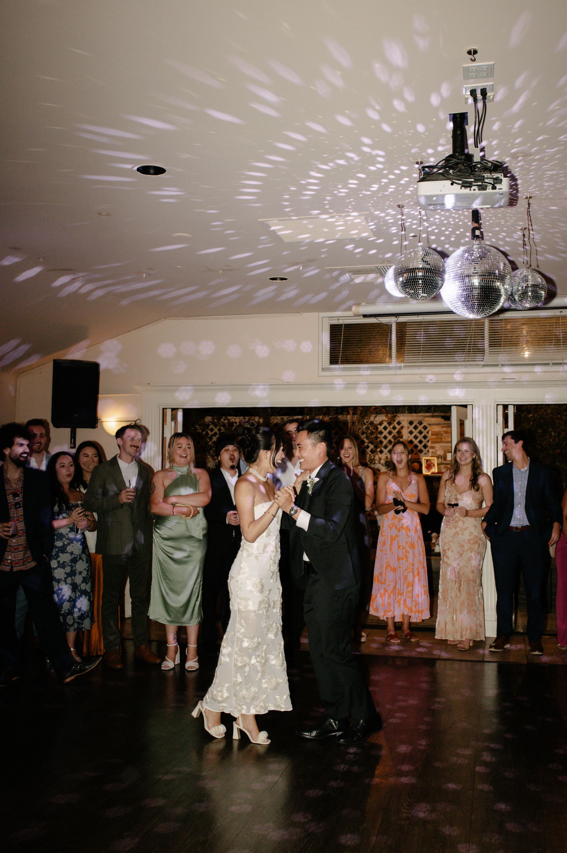 Bride and groom dance at a reception, with guests watching under sparkling ceiling lights.