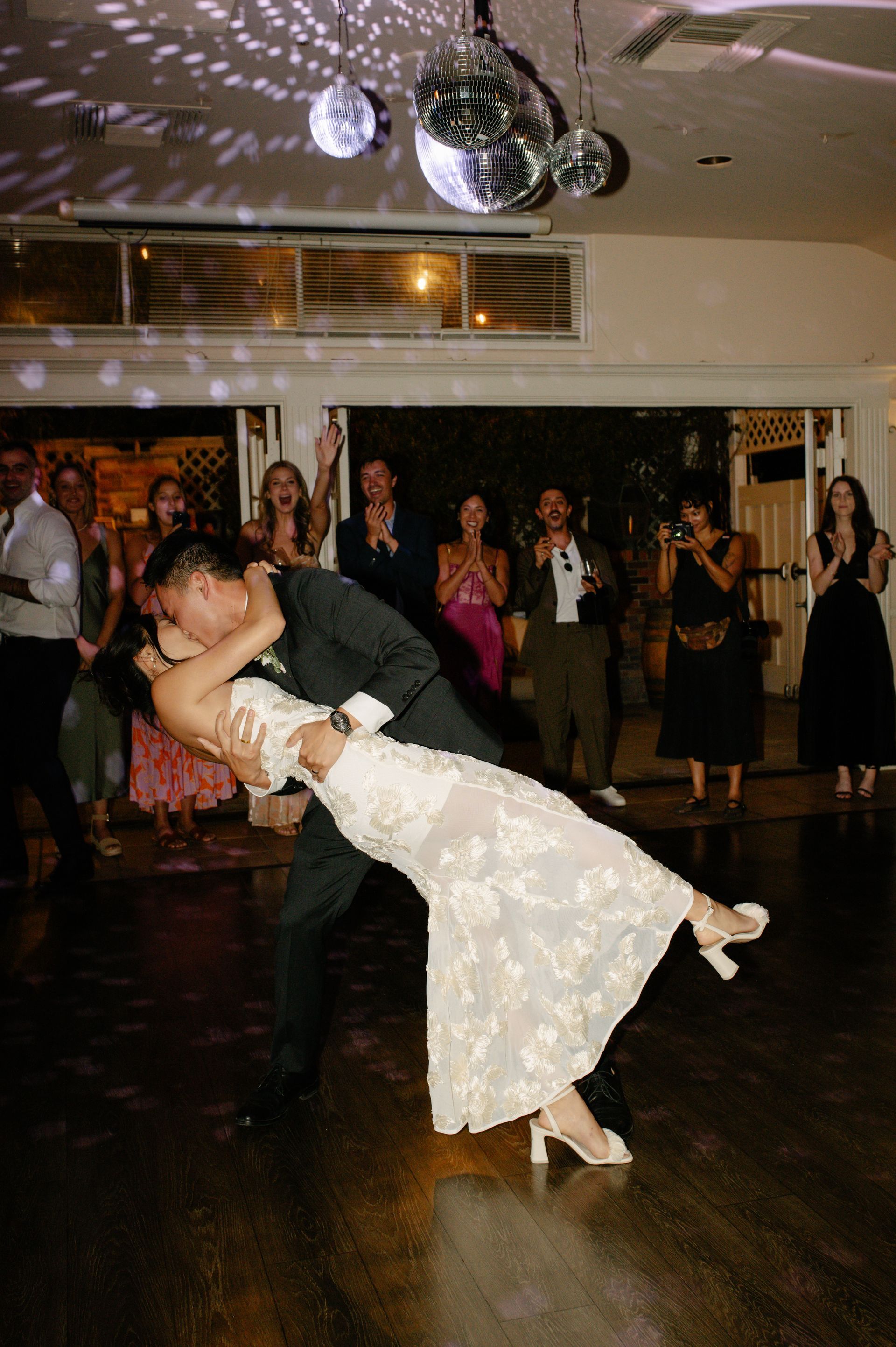 Couple dancing at a wedding reception on a dark wood floor, guests watching nearby