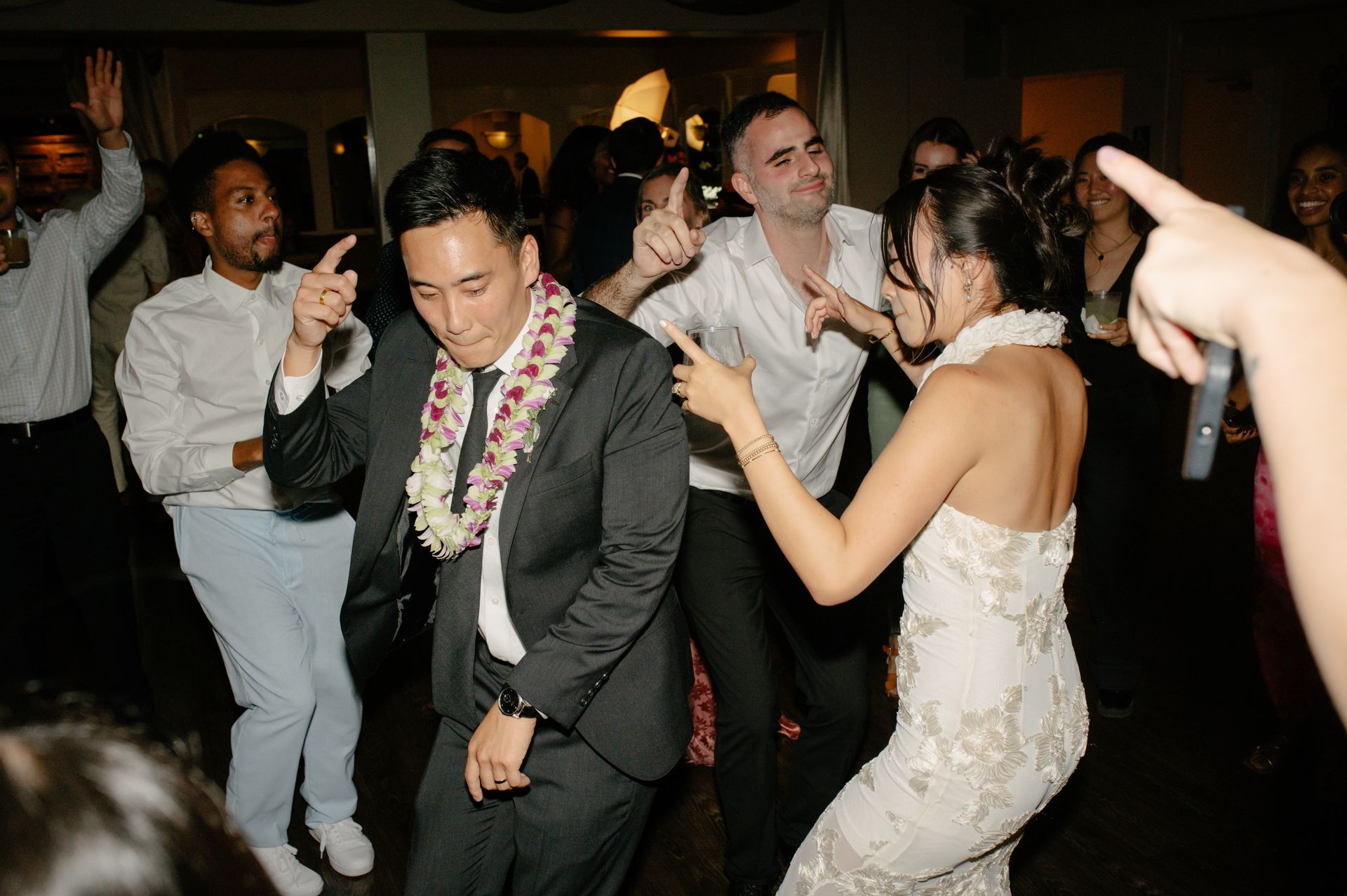 People dancing at a lively indoor party, with one man in a suit wearing a lei in the foreground.