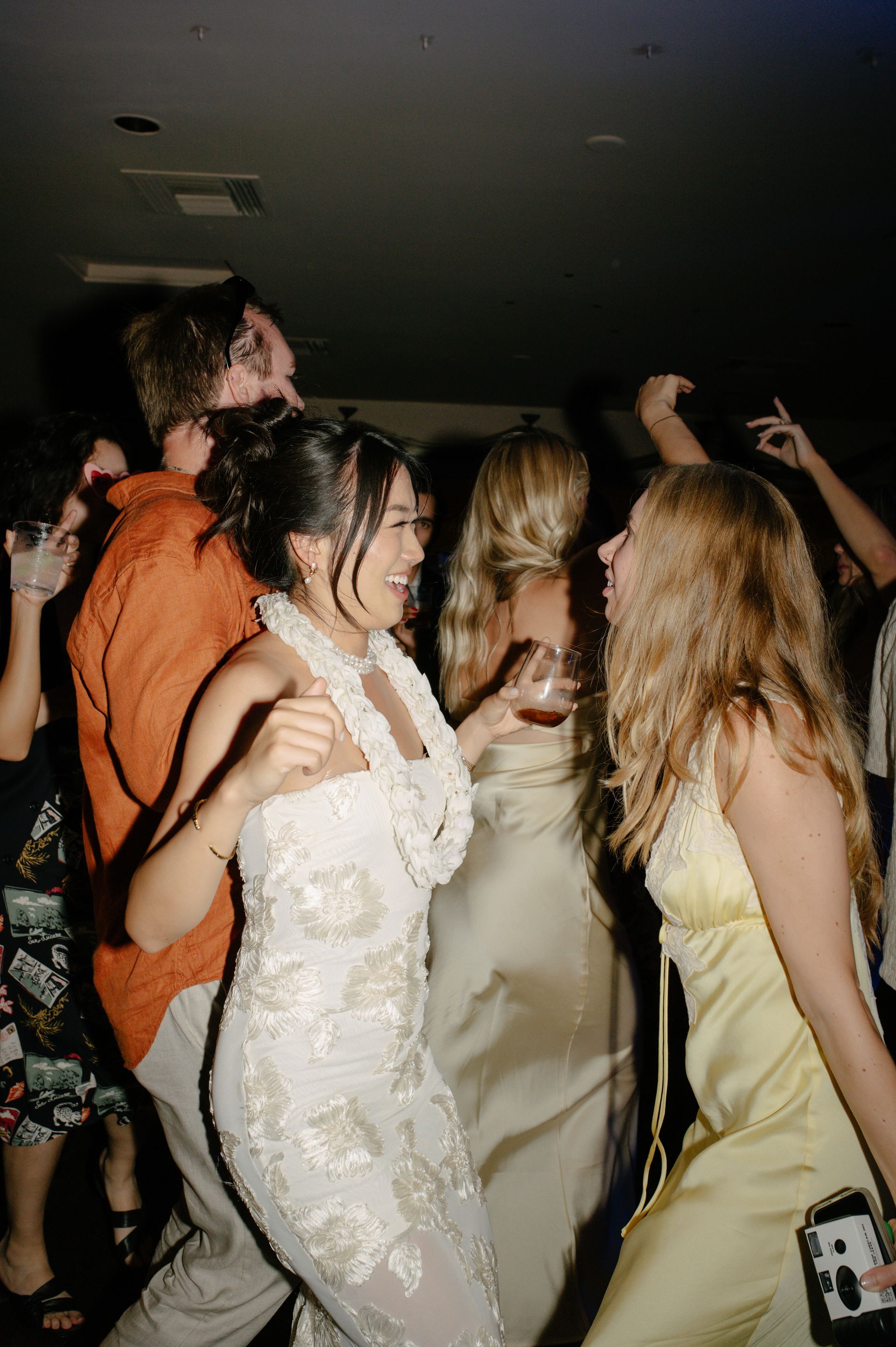 People dancing closely at a dim indoor party, with several wearing formal dresses and one person in orange.