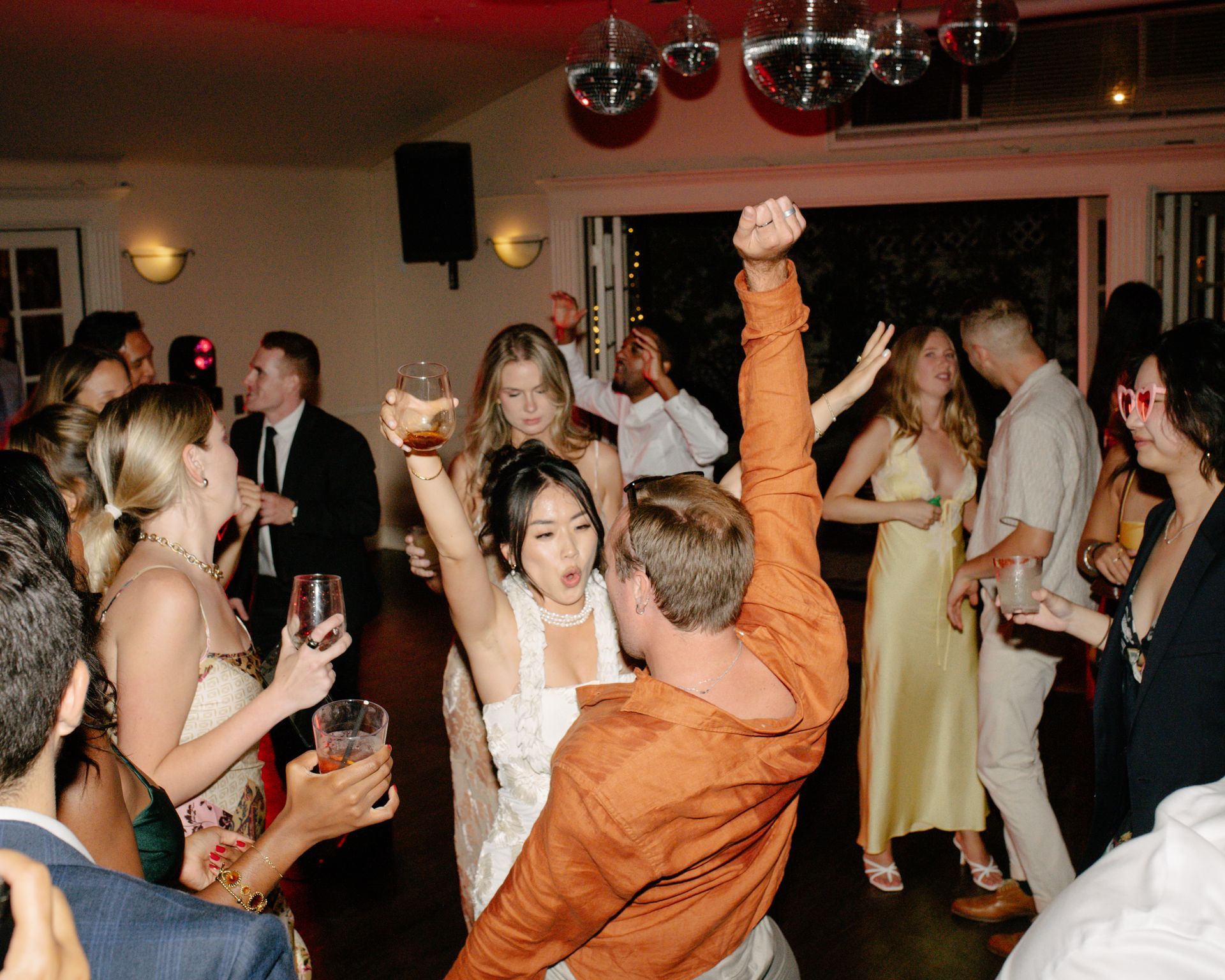 Crowded dance floor at a wedding reception, with guests dancing and holding drinks under warm indoor lighting