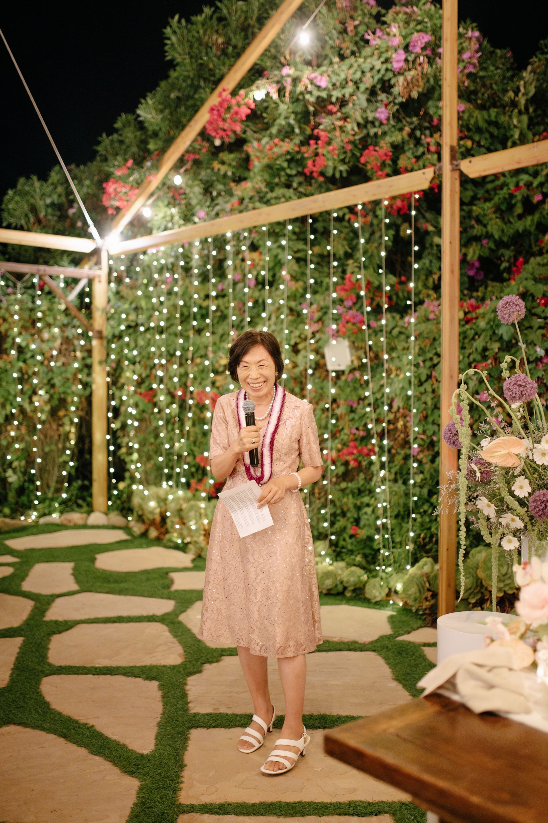 Woman standing on a flower-lit garden path at night, holding a bouquet under a decorated arch.
