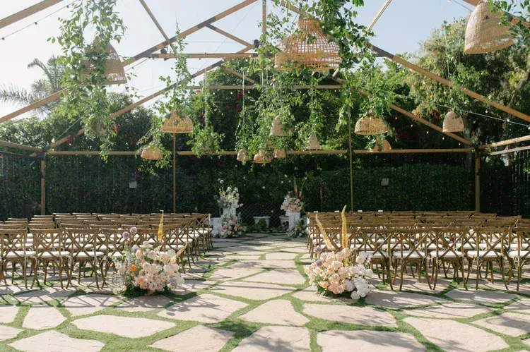 Outdoor wedding ceremony setup with flower-lined aisle, wooden chairs, and greenery under a canopy structure