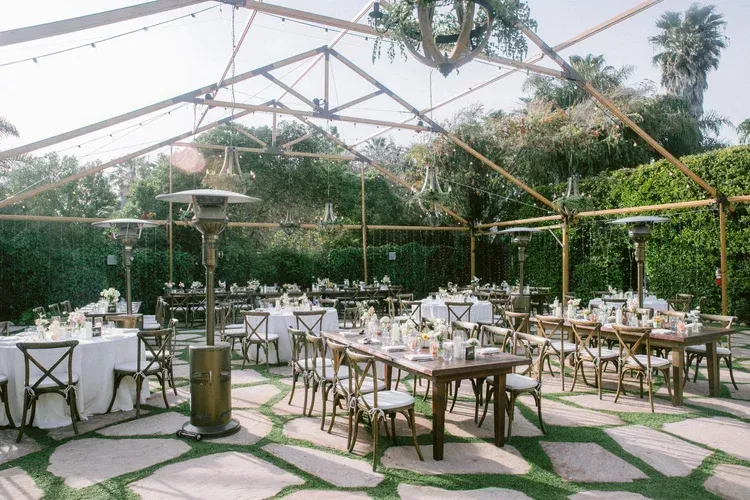 Outdoor event dining under a clear tent with white tables, gold chairs, and palm trees in the background