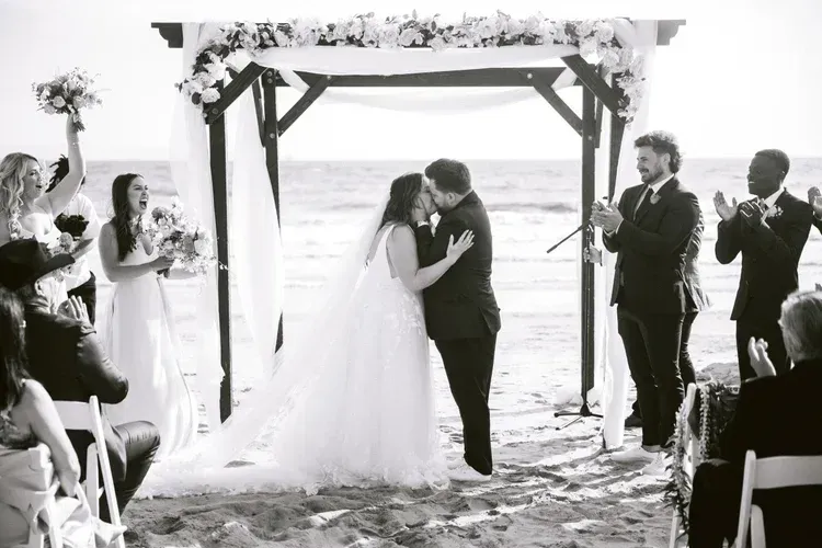 Bride and groom kiss under a beach wedding arch as guests applaud