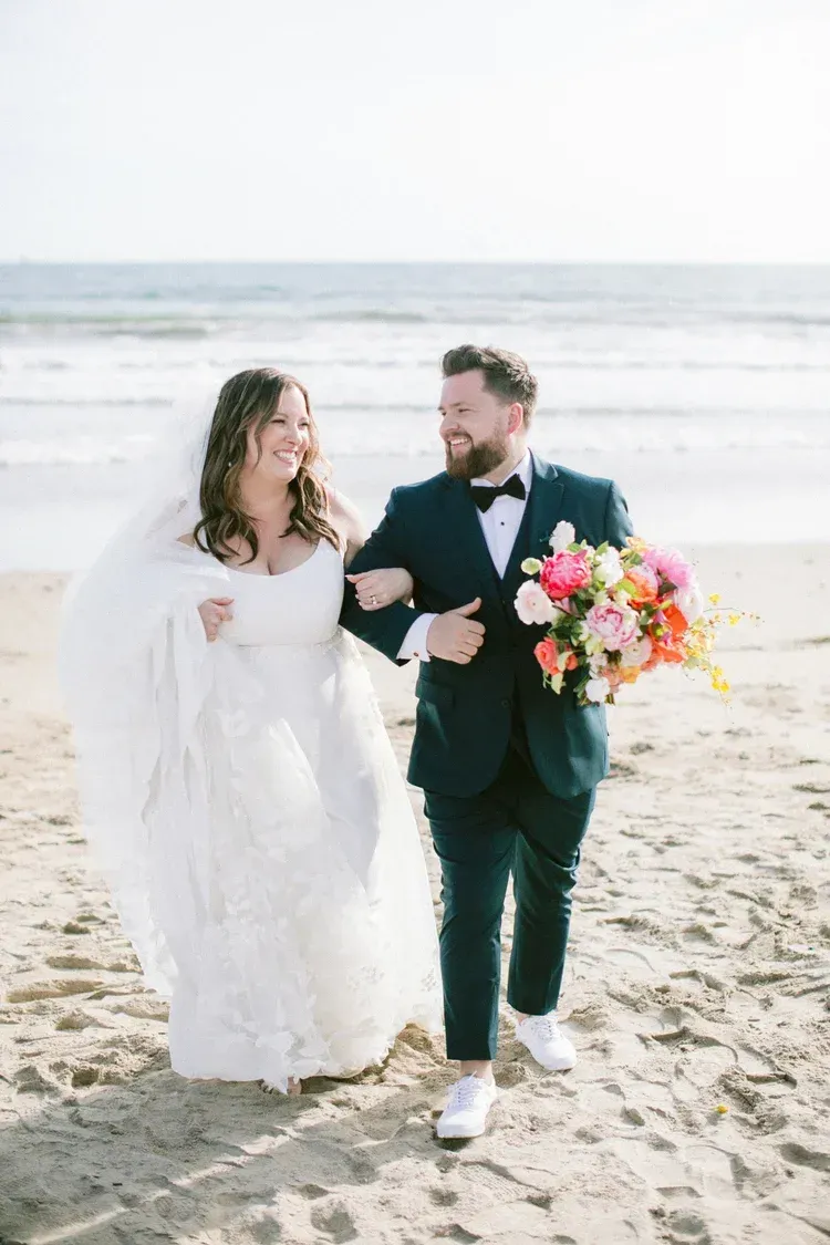 Newlyweds walking hand in hand on a beach, bride in white gown and groom in dark suit carrying a colorful bouquet