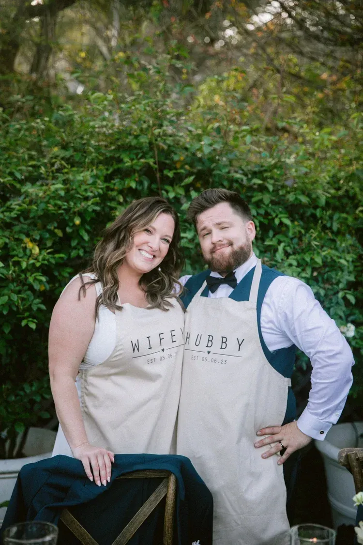 Two people smiling outdoors in aprons, standing behind a table with greenery in the background.