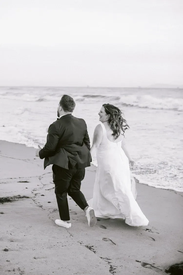 Two people holding hands and walking along a beach, with waves in the background.