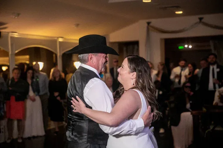 Couple dancing at a formal indoor event, with the man in a black hat and vest and the woman in a white dress.