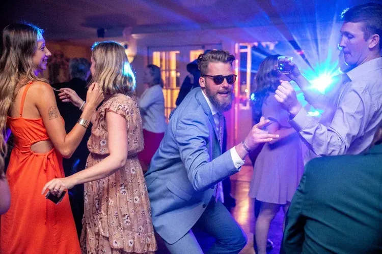 People dancing at a lively party with blue and purple lights, one man crouching in sunglasses.