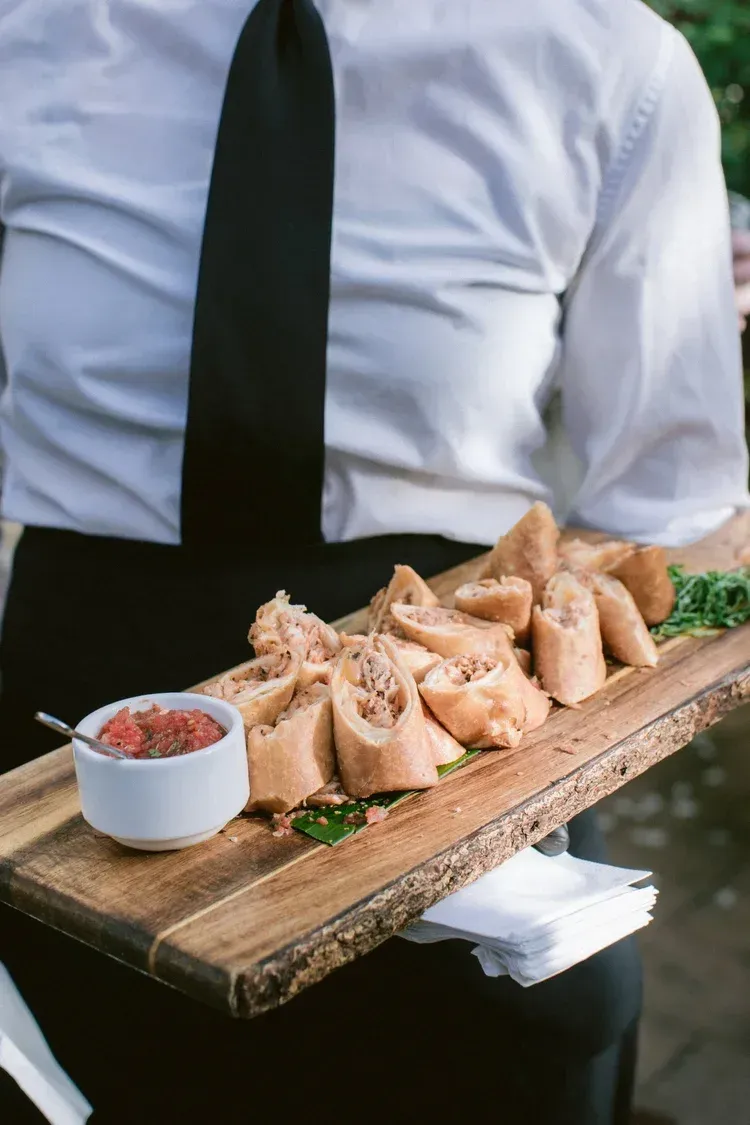 Waiter carrying a wooden tray of sliced bread with a small dipping sauce cup outdoors