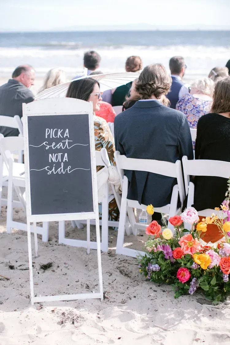 Beach wedding seating with white chairs, a chalkboard sign, and a colorful floral arrangement near the aisle