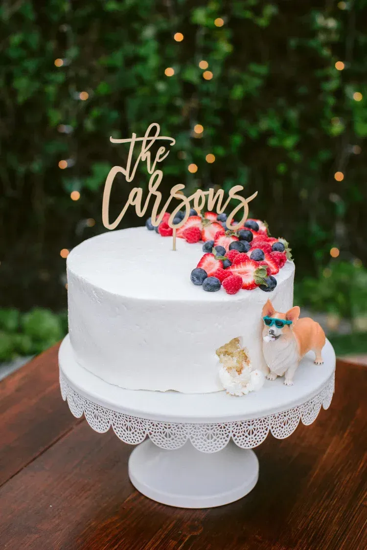 White decorated cake with berries and “the Carsons” topper on a lace stand outdoors