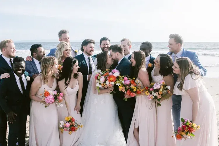 Wedding party in white and pastel attire posing outdoors with bouquets and smiling guests