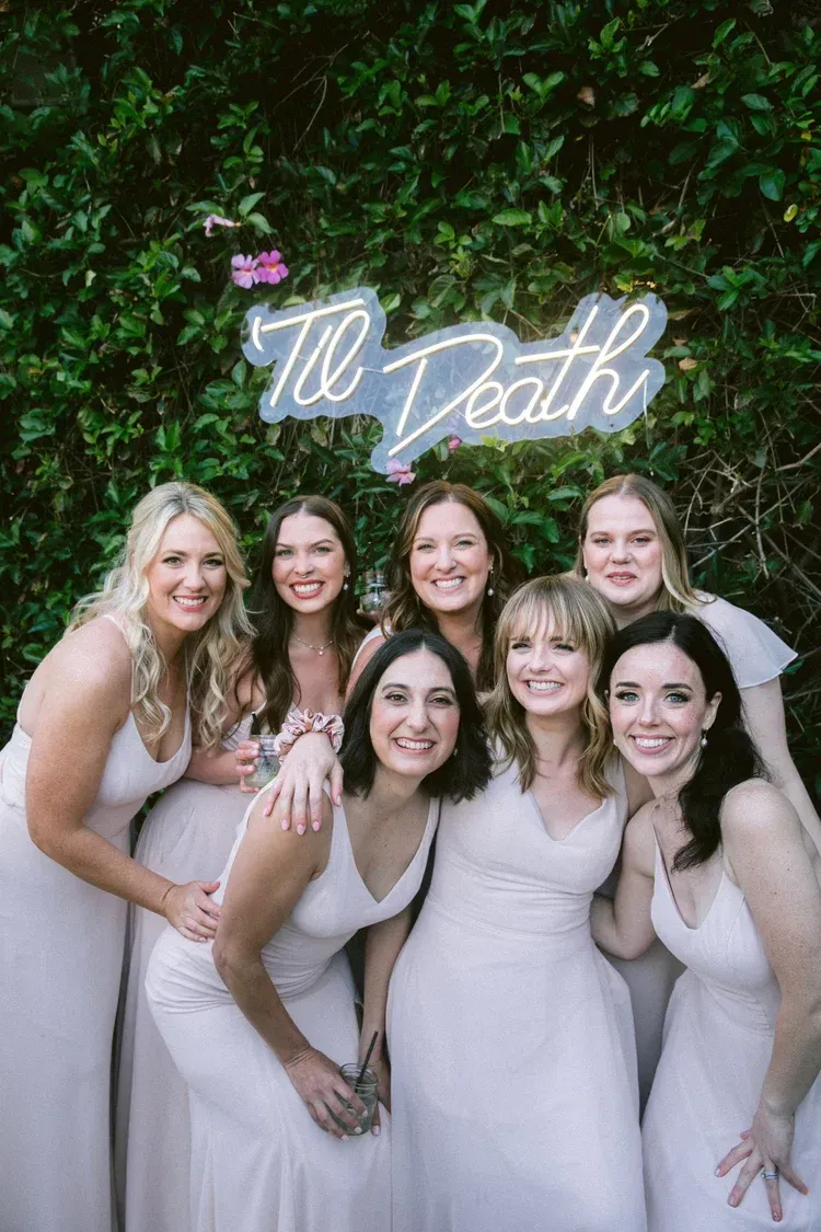 Six women in light dresses posing under a glowing “To Death” sign against a green leafy wall.