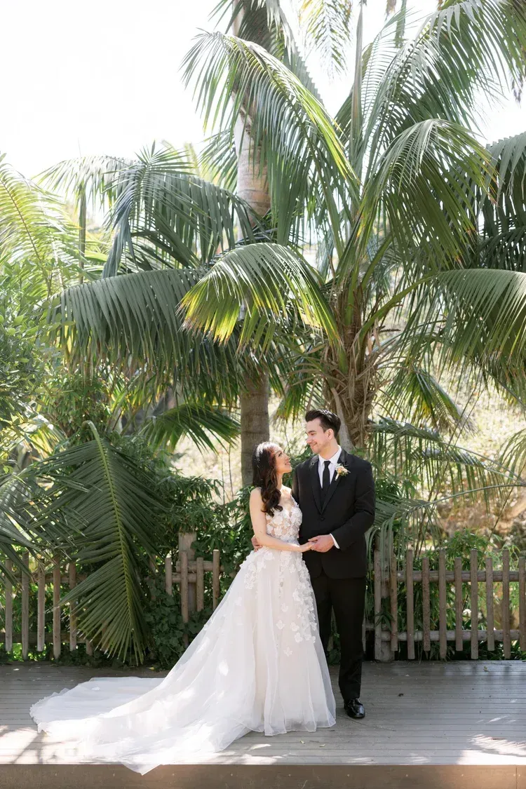 Bride and groom in white gown and black tuxedo, standing together outdoors beneath palm trees