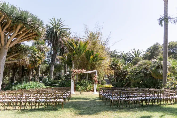 Rows of wooden chairs facing a floral arch in a sunny palm garden, set for an outdoor ceremony