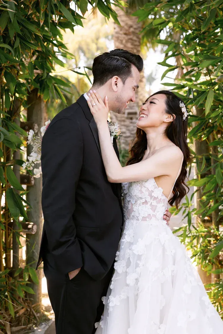 Bride and groom embracing under leafy greenery, gazing at each other outdoors in wedding attire