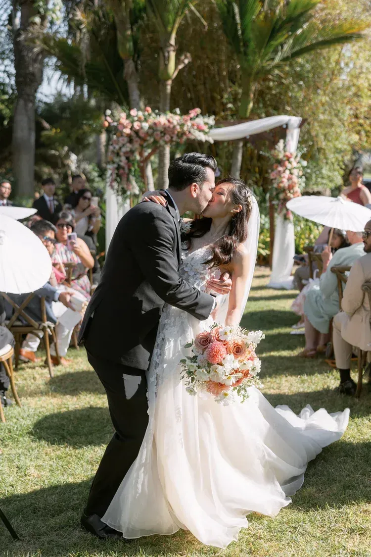 Bride and groom kiss at an outdoor garden wedding, with guests and palm trees in the background.