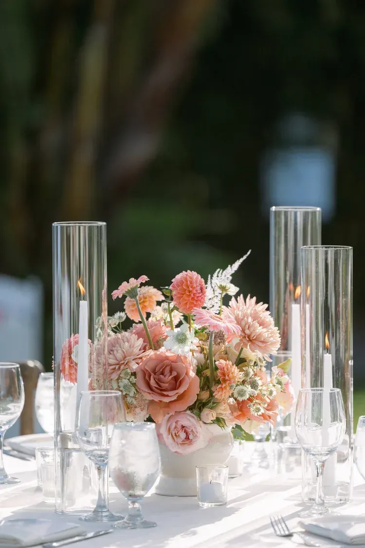 Elegant floral centerpiece with blush and peach roses on a white table, flanked by tall glass candle holders