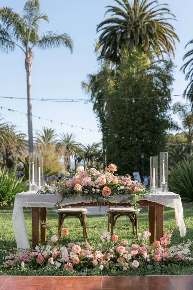Outdoor wedding table with pink floral arrangements and chairs on a lawn, with palm trees in the background