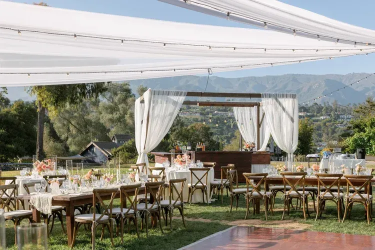 Outdoor wedding reception with long wooden tables, white drapes, and mountain views under a clear sky