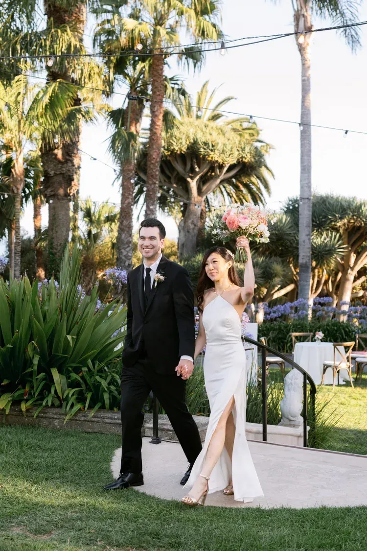 Bride and groom walk through a palm-lined garden, smiling and waving in formal wedding attire.