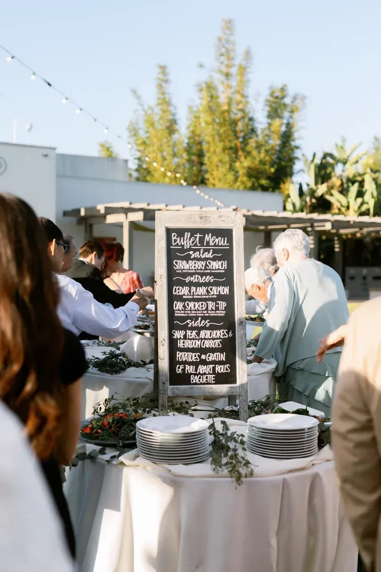 Outdoor buffet table with chalkboard menu and stacked plates at a sunny event