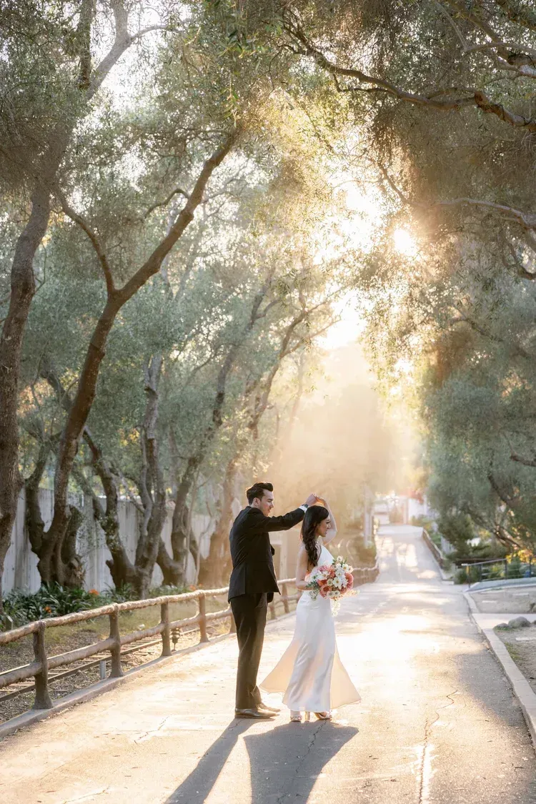 Couple posing on a sunlit tree-lined path, with soft golden light filtering through the branches.