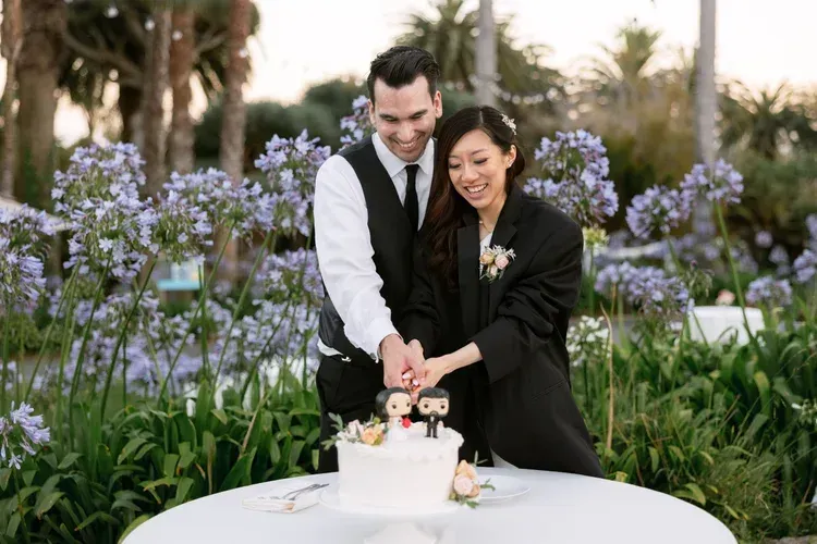 Couple cutting a small cake at an outdoor garden celebration with purple flowers