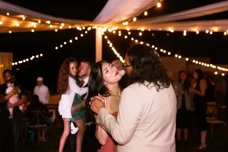 Couple dancing under string lights at an outdoor evening event, with guests in the background